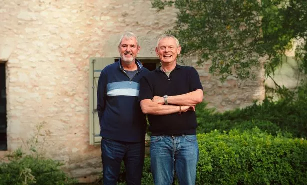 Neil Morrissey and Martin Clunes pose together outside a French cottage. Martin has his arms folded