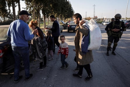 A woman and three small boys go to get into a car as an older man stands by and another man carries a large sack; they are watched by an armed fighter