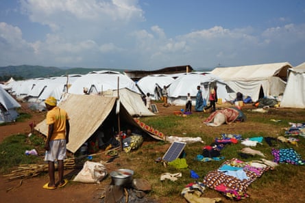 A person stands outside a tent in the foreground, with more tents in the background. Some possessions are scattered on the ground outside.