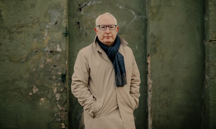 Neil Duncan-Jordan stands against a green, mossy wall in Poole. He is in his 60s and wears a black scarf and beige mac. 