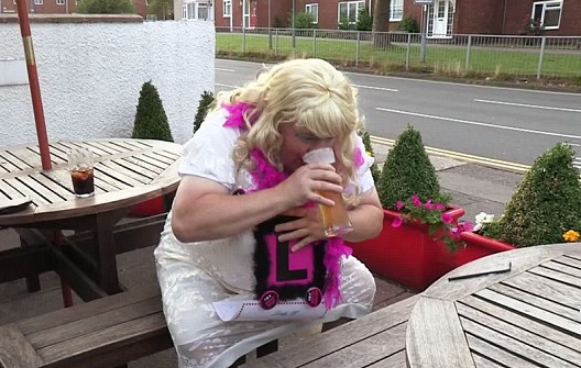 Person in a white dress and blonde wig drinking from a pint glass at an outdoor table.