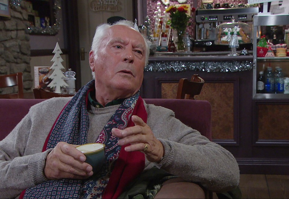 Anthony Miligan, an Emmerdale character, sits in a cafe, holding a cup.