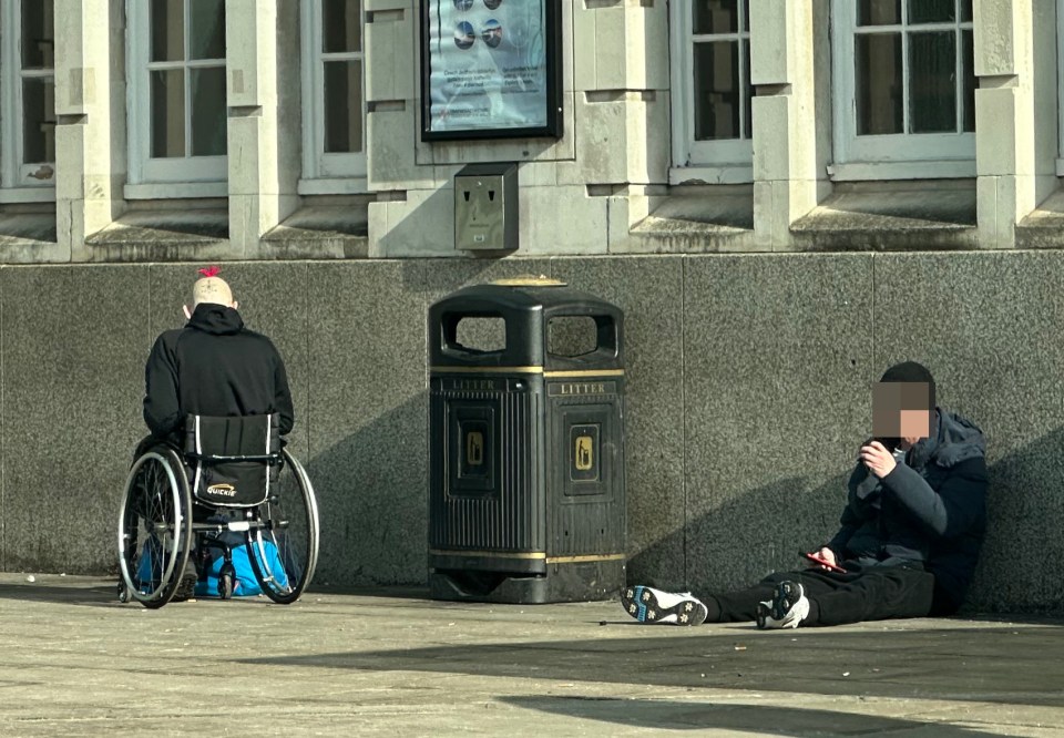 Two people, one in a wheelchair, sitting outside a railway station.