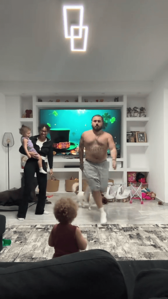 A family in their living room; a man is shirtless and dancing while a woman holds a young child.  Another young child watches.