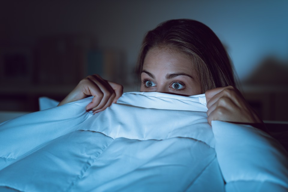 A frightened woman hides under her blanket while watching a scary movie.