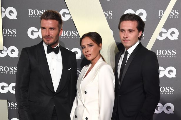 David Beckham, in a black suit, Victoria Beckham, in a white suit, and Brooklyn Beckham, in a black suit, at an awards ceremony in 2019.
