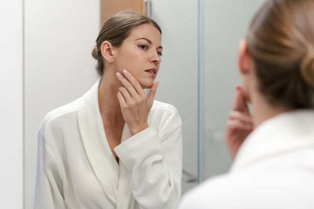 Young and gorgeous woman in white bathrobe standing in bright light bathroom with mirror. She looking at her reflection with calm face, holding hand on cheek, checking skin