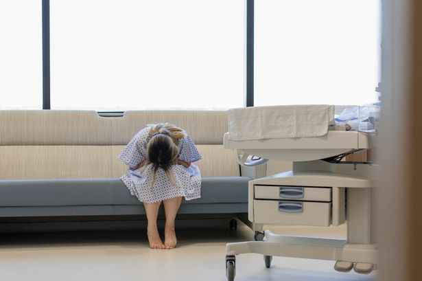 The young adult woman bends forward, puts her head in her lap and weeps in the hospital room. In the foreground is an infant warmer.