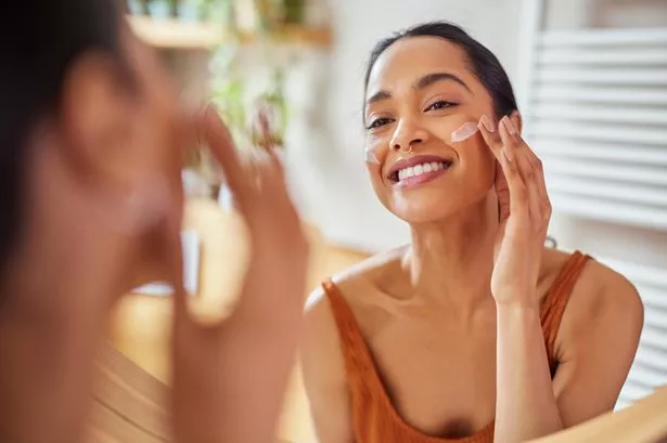 Portrait of smiling latin woman applying cream on her face while looking in mirror in her bathroom.