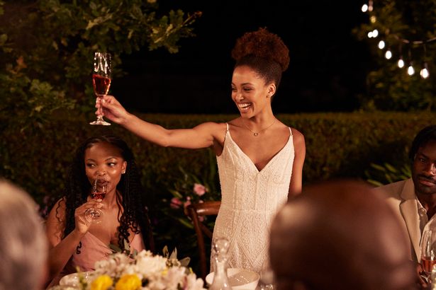 Cheerful young newlywed bride toasting drink while giving speech during wedding reception dinner party