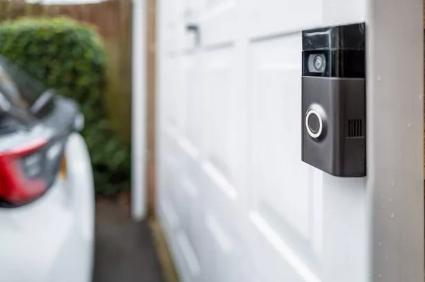 Shallow focus of a popular, battery powered wireless doorbell and integral camera seen located on a driveway, near a side gate entrance. A parked car is on the driveway.