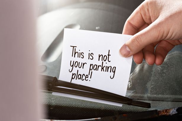 A hand places or removes a note from under the windscreen wiper of a car. It reads