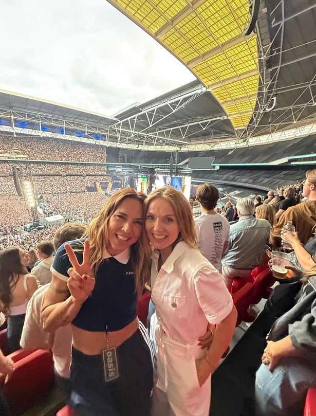 Mel C, in a blue top and trousers, stood beside Geri Horner, in a white jumpsuit, in the audience at Wembley Stadium.