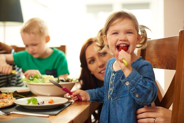 A young family of four sit at the dining table and eat pizza and salad. Mum helps her little girl with her meal , as she cheekily stuffs a whole tomato into her mouth