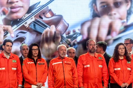 Politicians and executives wearing orange boiler-suits and appearing to be singing, stand in a line on a stage with a screen showing children playing violins behind them. 