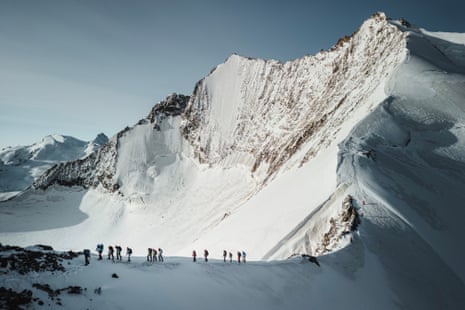 A group of hikers in the shadow of a sunlit, snowy mountain ridge