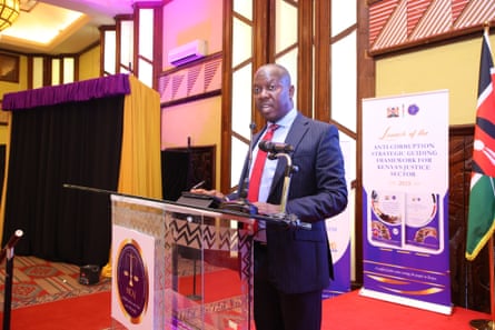 A middle-aged African man in a suit stands at a lectern, giving a speech