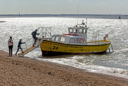 Foot passengers walking up a gangplank from a pebble beach to a small yellow ferry boat on a sunny day