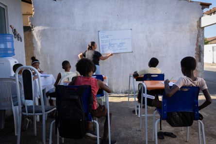 Five children sit at desks as a woman writes on a whiteboard on the wall of an open-air yard