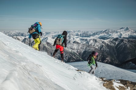 Three people walk down a snow-covered slope on a mountain with a view of the Alps in the background