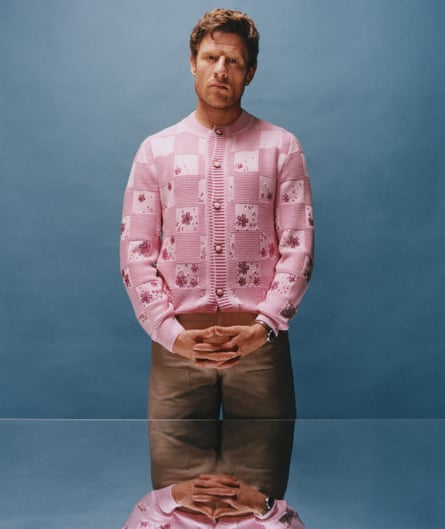 Actor James Norton, hands clasped below his waist, in front of a glass-topped table, in pink cardigan and brown trousers