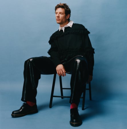 Actor James Norton, in black jumper and trousers, sitting on a chair, gazing to the side, against blue background