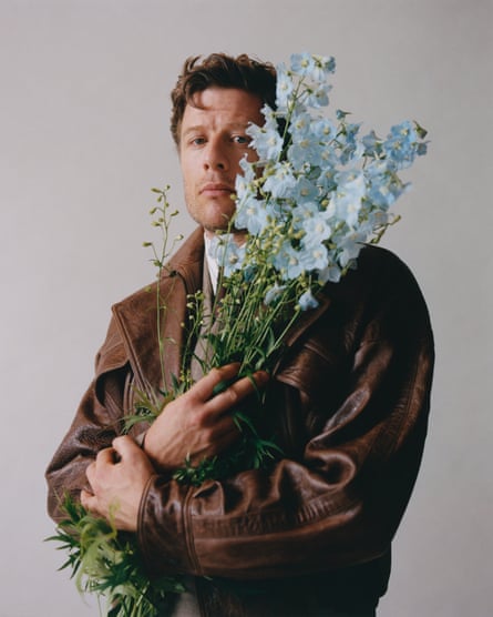 Actor James Norton, wearing a brown leather jacket and holding a bunch of tall pale blue flowers, July 2025