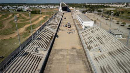 Two long concrete stands of seats on either side of a narrow arena space with a concrete arch in the background