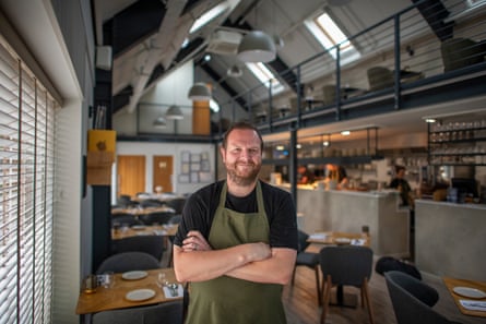 Dan Lawrence stands smiling in his restaurant with his arms folded: he has a gingery beard and wears a black top with khaki apron. The restaurant is in a barn-like building and is modern-looking with a high arched roof and upper layer of tables on a balcony over the bar and kitchen area