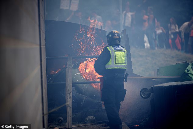 A police officer attempts to extinguish a wheelie bin that has been set alight by anti-migration protesters outside of the Holiday Inn Express