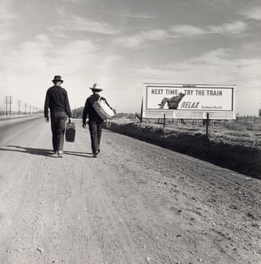 a black and white photo of two people walking along a road near a billboard that says ‘Next time, try the train. Relax’