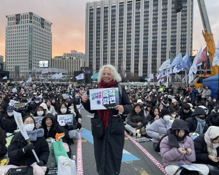 Standing older white woman bearing a placard in South Korean with many hundreds of young South Koreans seated behind her peacefully protesting.