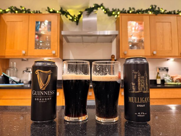 A can of Guinness, with its contents poured into a pint glass, next to a can of Mulligan's, which has also been poured into a pint glass. The cans and glasses are on a kitchen work surface