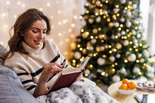 Portrait of happy young woman lounging on the cozy sofa, under a warm blanket, reading an interesting book. Christmas tree and lights glistening in the background.