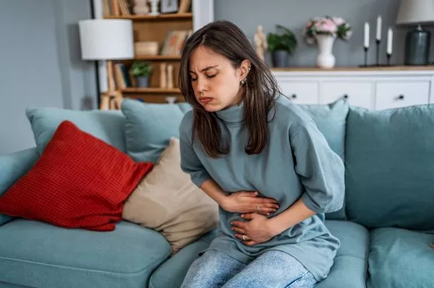 Young woman sitting on a sofa at home, experiencing abdominal pain and discomfort, holding her stomach with a worried expression