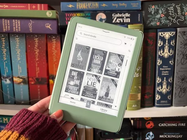 A hand holding the matcha Kindle in front of a bookcase 