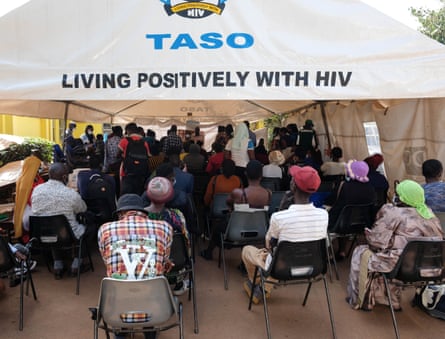 A few dozen African people seen from behind as they sit in a waiting area under a tent