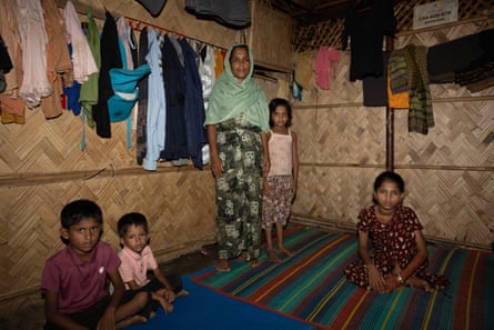 A woman stands on a mat inside a shelter made of woven bamboo. Children sit near her and clothing hangs from the walls.